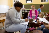 Vista lateral de una adolescente afroamericana en una clase de secundaria sentada en un escritorio, concentrándose y escribiendo, con compañeros de clase adolescentes masculinos y femeninos sentados en escritorios trabajando en segundo plano - foto de stock