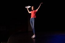 Vista laterale di un'adolescente caucasica del liceo in un teatro vuoto del liceo che si prepara prima di uno spettacolo, in piedi sul palco con le braccia in aria, con un copione in mano e praticando la sua parte — Foto stock