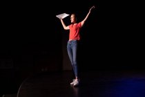 Side view of a Caucasian high school teenage girl in an empty high school theatre preparing before a performance, standing on the stage with arms in the air, holding a script and practicing her part — Stock Photo