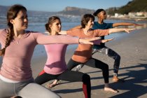 Vista lateral de un grupo multiétnico de amigas disfrutando de hacer ejercicio en una playa en un día soleado, practicando yoga de pie en la asana guerrera. - foto de stock