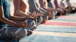 Вид сбоку на середину группы подруг, наслаждающихся упражнениями на пляже в солнечный день, практикующих йогу, сидящих в позе йоги с руками-мудрой, посредничающих. — стоковое фото