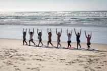 Вид спереду на багатоетнічну групу друзів-жінок, які насолоджуються фізичними вправами на пляжі в сонячний день, практикуючи йогу, стоячи в позі йоги . — стокове фото