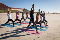 Вид збоку на багатоетнічну групу друзів, які насолоджуються фізичними вправами на пляжі в сонячний день, практикують йогу, стоячи в позі йоги . — стокове фото