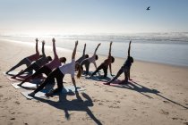 Rückansicht einer multiethnischen Gruppe von Freundinnen, die an einem sonnigen Tag am Strand Sport treiben, Yoga praktizieren, sich in Yogaposition dehnen. — Stockfoto