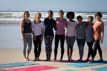 Вид спереди многоэтнической группы подруг, веселящихся вместе на пляже в солнечный день, стоящих за ковриками для йоги, одетых в спортивную одежду, улыбающихся, улыбающихся.. — стоковое фото