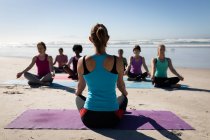 Vue arrière de la femme caucasienne, vêtue de vêtements de sport, assise sur un tapis de yoga, pratiquant le yoga avec un groupe d'amis féminines multi-ethniques assis face à elle sur la plage ensoleillée. — Photo de stock