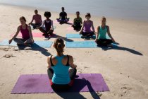 Rückansicht einer kaukasischen Frau, die Sportkleidung trägt, auf einer Yogamatte sitzt und mit einer Gruppe multiethnischer Freundinnen Yoga praktiziert, die ihr am sonnigen Strand gegenübersitzen. — Stockfoto