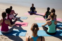 Vista trasera de un grupo multiétnico de amigas disfrutando de hacer ejercicio en una playa en un día soleado, practicar yoga, sentarse en posición de yoga, meditar. - foto de stock