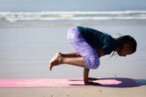 Vista laterale di una donna afroamericana attraente, indossa vestiti sportivi, pratica yoga, in piedi nelle sue mani in posizione yoga, sulla spiaggia soleggiata. — Foto stock