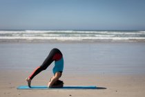 Vista laterale di una donna attraente caucasica, indossando vestiti sportivi, praticando yoga, in piedi in posizione yoga, preparandosi alla testata, sulla spiaggia soleggiata. — Foto stock