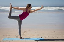 Seitenansicht einer kaukasischen attraktiven Frau, die Sportkleidung trägt, Yoga praktiziert, mit einem Bein in Yogaposition am sonnigen Strand steht. — Stockfoto