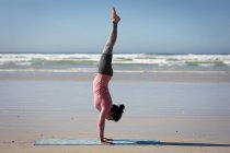 Vue latérale d'une femme séduisante caucasienne, portant des vêtements de sport, pratiquant le yoga, debout dans ses mains en position de yoga, sur la plage ensoleillée. — Photo de stock