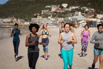 Vista frontal de un grupo multiétnico de amigas disfrutando de hacer ejercicio en una playa en un día soleado, corriendo a la orilla del mar. - foto de stock