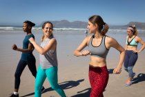 Vista lateral de un grupo multiétnico de amigas disfrutando de hacer ejercicio en una playa en un día soleado, corriendo a la orilla del mar. - foto de stock
