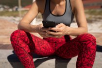 Vista frontal sección media de una mujer, con ropa deportiva, sentado en la orilla de la playa, relajarse después de trotar, sosteniendo su teléfono inteligente en ambas manos, masaje de mensajes de texto. - foto de stock