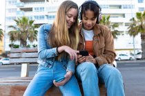 Front view of a Caucasian and a mixed race girls enjoying time hanging out together on a sunny day, sitting on a bench, girl wearing headphones, holding smartphone and showing it to her friend. — Stock Photo