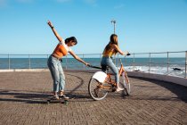 Vista lateral de un caucásico y una mezcla de chicas de carreras disfrutando del tiempo juntos en un día soleado, jugando en el paseo marítimo, una chica montando una bicicleta y tirando de otra en el monopatín. - foto de stock