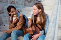 Vue de face d'un Caucasien et une mixte filles profiter du temps passer ensemble par une journée ensoleillée, manger de la crème glacée, assis sur les escaliers dans une promenade au bord de la mer. — Photo de stock