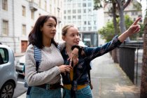 Передній вид на кавказьку і змішану расу дівчат, які проводять час разом в сонячний день, стоячи на тротуарі, обіймаючи, дівчинка вказує. — стокове фото