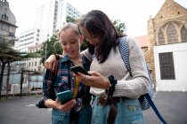 Передній вид на кавказьку і змішану расу дівчат, які проводять час разом у сонячний день, стоять на тротуарі, обіймають, тримаючи свої смартфони. — стокове фото