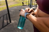 Vista lateral sección media de una mujer deportiva haciendo ejercicio en un gimnasio al aire libre durante el día, usando su reloj inteligente, sosteniendo una botella de agua. - foto de stock