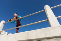 Niedrige Seitenansicht einer sportlichen kaukasischen Frau mit langen dunklen Haaren, die an einem sonnigen Tag mit blauem Himmel auf einer Strandpromenade trainiert, ihr Bein ausstreckt und sich an ein Geländer lehnt. — Stockfoto