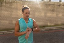 Вид спереди спортивной белой женщины с длинными темными волосами, тренирующейся в городской местности в солнечный день, проверяющей свои умные часы. — стоковое фото