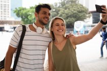Vista frontal de una feliz pareja caucásica por las calles de la ciudad durante el día, parada en la calle y tomando una selfie con su teléfono inteligente. - foto de stock