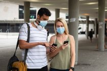 Vista frontal de una pareja caucásica por las calles de la ciudad durante el día, con máscaras faciales contra la contaminación del aire y covid19 coronavirus, utilizando sus teléfonos inteligentes. - foto de stock
