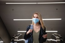 Вид белой женщины с длинными слепыми волосами, покидающей эскалатор, использующей смартфон и маску для лица против загрязнения воздуха и вируса коронавируса covid19. — стоковое фото