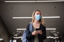 Vorderansicht einer kaukasischen Frau mit langen, blinden Haaren, die eine Rolltreppe verlässt, ihr Smartphone benutzt und eine Gesichtsmaske gegen Luftverschmutzung und Covid19 Coronavirus trägt. — Stockfoto