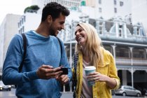 Vista frontal de una feliz pareja caucásica por las calles de la ciudad durante el día, sosteniendo una taza de café para llevar, usando un teléfono inteligente y sonriendo. - foto de stock