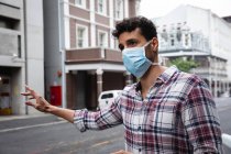 Vista frontal close-up de um homem caucasiano vestindo camisa quadriculada e máscara facial contra a poluição do ar e covid19 coronavírus, saudando um táxi na rua . — Fotografia de Stock