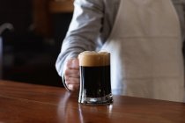 Una mezza sezione di uomo che lavora in un pub di microbirreria, indossa un grembiule bianco, serve una pinta di birra, la mette su un bar di legno. — Foto stock