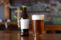 Ein Glas echten Bieres mit einem Schaumkopf und einer Glasflasche sitzt auf der Holztheke einer Mikrobrauerei. — Stockfoto