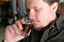 Close up of a Caucasian man with long hair working in a microbrewery, holding a glass of beer, smelling it for inspection. — Stock Photo