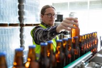 Hombre caucásico con chaleco de alta visibilidad, trabajando en una microcervecería, comprobando botellas de vidrio oscuro de cerveza en una cinta transportadora. - foto de stock