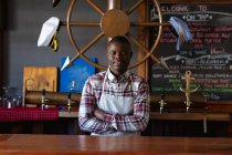 Portrait d'un barman afro-américain portant un tablier blanc, travaillant dans un pub de microbrasserie, les bras croisés et regardant droit dans une caméra. — Photo de stock