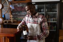 Uomo afroamericano che lavora in un pub di microbirreria, indossa un grembiule bianco, tiene una bottiglia di birra e guarda dritto in una macchina fotografica. — Foto stock