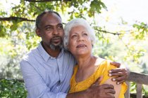 Retrato de um casal afro-americano sênior desfrutando de sua aposentadoria, sentado em um jardim ao sol abraçando e olhando para o lado sorrindo, casal isolando durante a pandemia do coronavírus covid19 — Fotografia de Stock