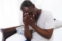 Крупным планом красивый пенсионер афроамериканец сидит дома на кровати с головной болью, держа голову в боли и глядя вниз,, самоизоляция во время коронавируса covid19 пандемии — стоковое фото