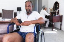 A senior retired African American couple at home in their bedroom, the man sitting in a wheelchair in his underwear using a smartphone and the woman sitting at her dressing table in the background, self isolating during coronavirus covid19 pandemic — Stock Photo