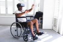 Un anciano afroamericano jubilado en casa, sentado en una silla de ruedas en ropa interior frente a una ventana en un día soleado usando un casco VR con los brazos extendidos frente a él, auto aislándose durante la pandemia de coronavirus covid19 - foto de stock