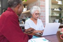 Una pareja de ancianos afroamericanos jubilados sentados en una mesa en su comedor, mirando el papeleo y discutiendo sus finanzas, la mujer usando una computadora portátil, en casa juntos aislándose durante la pandemia de coronavirus covid19 - foto de stock