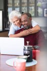 Close up of a happy senior retired African American couple at a table in their dining room, using a laptop computer together, the man sitting and the woman standing behind and embracing him, both smiling — Stock Photo