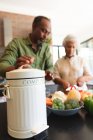 Занятая пожилая афроамериканская пара дома, готовит еду, режет овощи, кладет отходы в компостный контейнер на кухне, дома вместе изолирует во время пандемии коронавируса 19 — стоковое фото