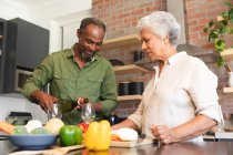 Glückliche Senior-Rentner afroamerikanische Paar zu Hause, die Zubereitung von Gemüse, um eine Mahlzeit zu machen, und der Mann schenkt ihnen Gläser Wein, Paar zu Hause zusammen Isolierung während Coronavirus covid19 Pandemie — Stockfoto
