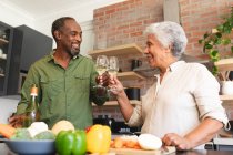 Happy senior aposentado casal afro-americano em casa, preparando legumes para fazer uma refeição, e fazer um brinde com copos de vinho branco, casal em casa juntos isolando durante coronavírus covid19 pandemia — Fotografia de Stock