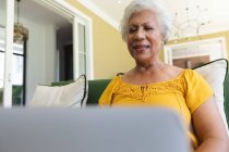 Nahaufnahme einer glücklichen attraktiven älteren afroamerikanischen Rentnerin, die zu Hause in einem Sessel in ihrem Wohnzimmer sitzt, einen Laptop benutzt und während der Coronavirus-Pandemie lächelt, sich selbst isoliert 19 — Stockfoto