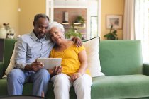 Happy senior aposentado casal afro-americano em casa sentado em um sofá em sua sala de estar, abraçando e usando um computador tablet juntos e sorrindo, casal isolando durante coronavírus covid19 pandemia — Fotografia de Stock